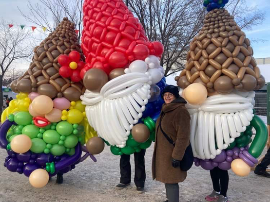 Senator Simons, in a brown wool coat and blue hat, grins as she poses with three performers in gnome suits made out of dozens of balloons.
