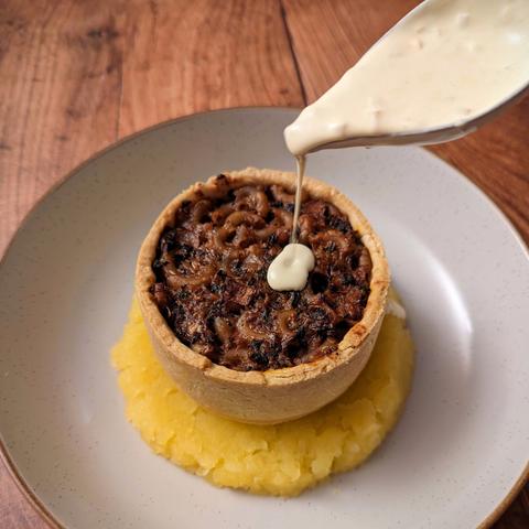 An individual open-top pie filled with a dark brown-red filling of macaroni and haggis, sitting on a circle of mashed neeps. A spoon is pouring a creamy sauce on top of the pie. It sits in a plate on a dark wood background.