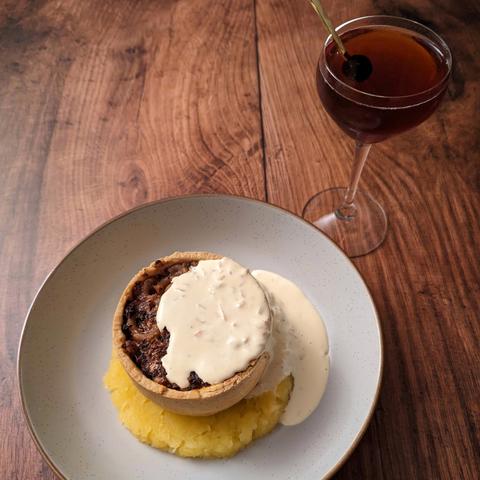 An individual open-top pie filled with a dark brown-red filling of macaroni and haggis, sitting on a circle of mashed neeps, with a creamy sauce poured over the top and pooling off one side. It is sat next to a dark red-brown cocktail with a maraschino cherry garnish in a Nick and Nora. Both sit on a brown wood backgound.