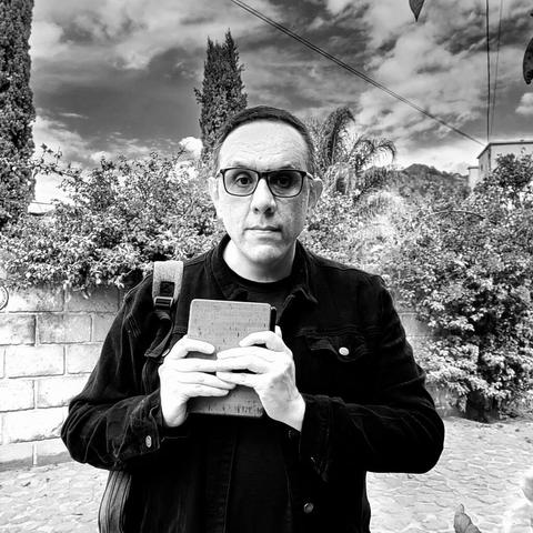 Delante de un cielo con nubes y de una barda de ladrillo bordeada por árboles, un hombre de lentes y vestido de hecho sostiene un lector de libros electrónicos entre las manos.