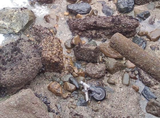Part of a scooter lying on rocks at the base of a cliff with salt water waves around it.