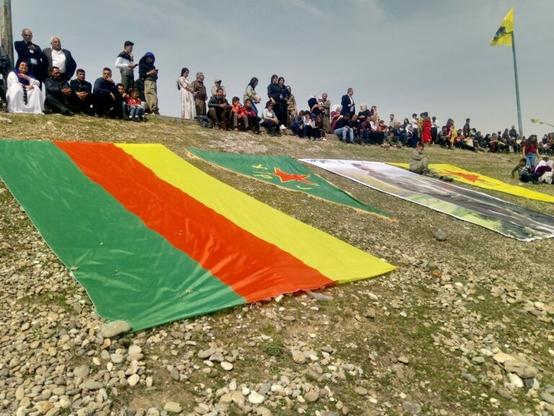 A photograph of a hill with various Kurdish flags and emblems on it with people gathered at the top of the hill.