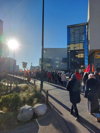 Un cortège défile contre la loi immigration dans la rue qui longe la gare à Annecy. Un beau soleil d'hiver illumine la manifestation, on voit des drapeaux syndicaux.