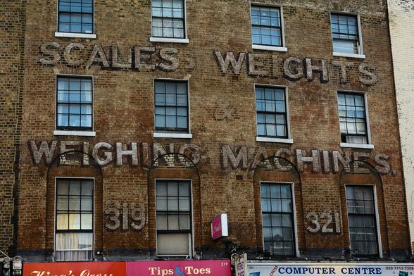 Top three floors of a large London building with fading painted letters that read "Scales, Weights, & Weighing Machines. 319, 321." where the numbers correspond to the two premises that the business occupied.