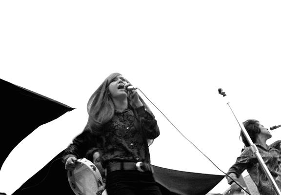 A low angle photo of Mary Weiss singing on an outdoor platform and overcast afternoon. Two black rectangular canvas rain shields flap above and behind her. She hold a tambourine in her lowered right hand, and sings into a microphone she holds close to her mouth in her left. Weiss’s mouth is wide and eyes narrowed. Another singer and tambourine player stands and sings to her left.