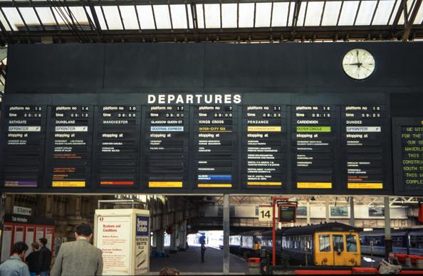 Solari departures board at Edinburgh Waverley in 1989, showing information for eight trains, including platform number, time, destination, train type, stopping points and onboard facilities. An old blue/grey BR class 108 DMU sits in platform 15 just behind the board.