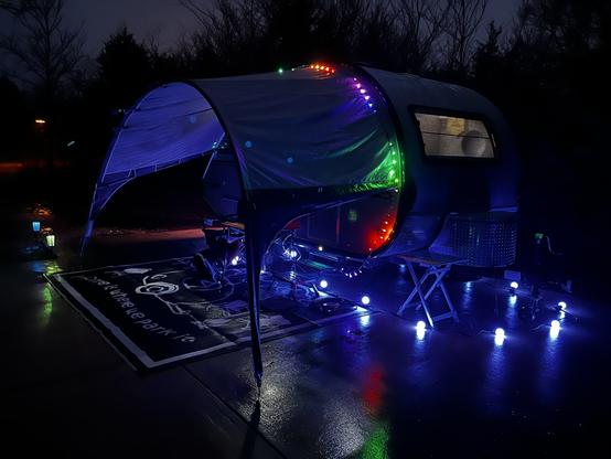 Clam shell shaped travel trailer with awning and festive lights. Picture is taken at night time.