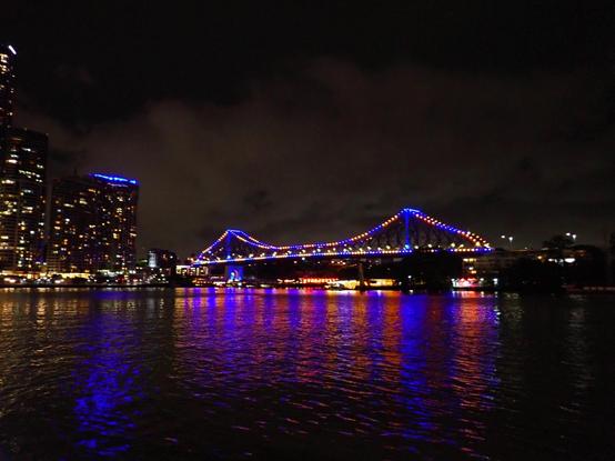 View of suspension bridge lit up at night in blues and reds, reflecting on the rippling river below