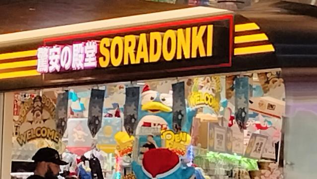 Japanese storefront for a mini Don Quijote in Haneda Airport. There are various spinner racks, machines with flashing lights, a sign in Japanese and English in yellow and white letters, and the blue penguin mascot is visible through the door.