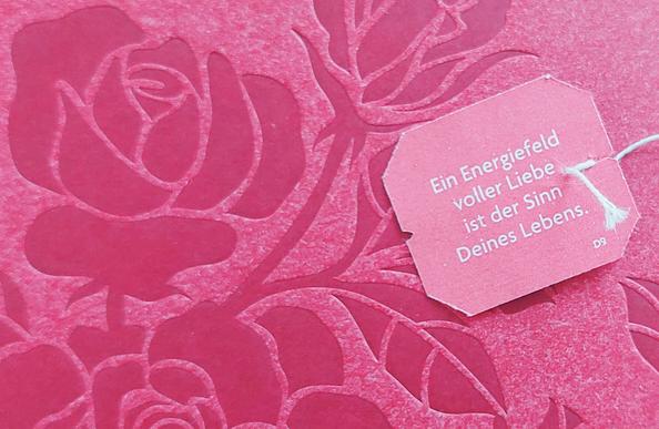 Auf einem roten Bild mit verschieden weit geöffneten Rosenblüten liegt ein Teebeutel-Anhänger, auf dem steht: "Ein Energiefeld voller Liebe ist der Sinn deines Lebens."