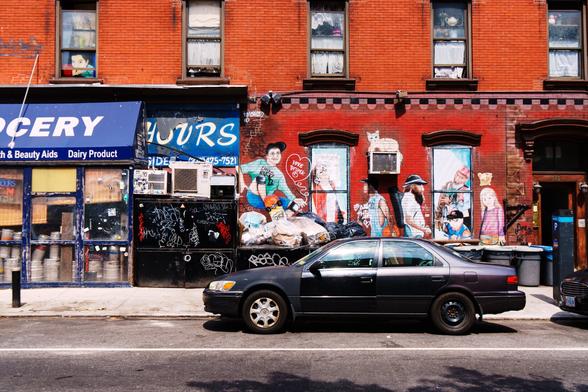 A color photograph of a brick building. A bodega occupies the lower level, and the blue awning stretches to the left out of the frame. Art is painted on the side, showing various people, including a skateboarder, saxophone player, basketball player, etc.  Another image of a face sits in a 2nd floor window to the left. Curtains obscure the others, but some items like a teapot sit on shelves above the curtains in the windows to the right. Metal sheds, dumpsters, and trash barrels with bags sitting on top line the sidewalk along the building. A somewhat banged up dark blue sedan, every body panel and door reflecting light differently, is parked in front.