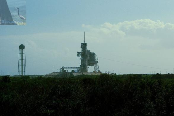 An outdoor, daylight photograph. The bottom third of the frame is dominated by green trees. At the horizon, a metal truss structure stands with a single, tall tower capped by a white cylinder and a wider section to the right of the tower that is almost rectangular in shape. A tall water tower is off to the far left, and the cloudy sky takes up the background.