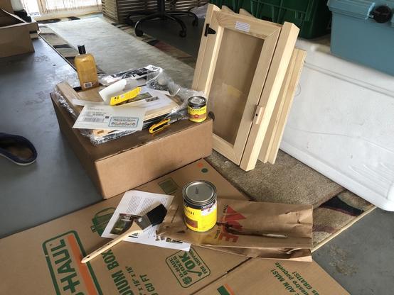 photo: daytime inside a garage. Assorted wood panels, a glass paned door, all to "large birdhouse" scale. Cardboard on the floor to work on. Can of wood stain and a paintbrush.
