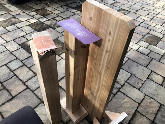 photo: driveway, outdoors and daytime, some larger wood pieces (the mounting post and an unassembled bench accessory) and some sandpaper of various grit.