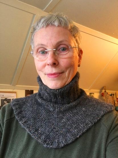 Older woman wearing a knitted neck warmer (collar & yoke) in a fuzzy soft tweedy grey yarn. It has a ribbed turtleneck and extends down to over the collarbones to the shoulders.
