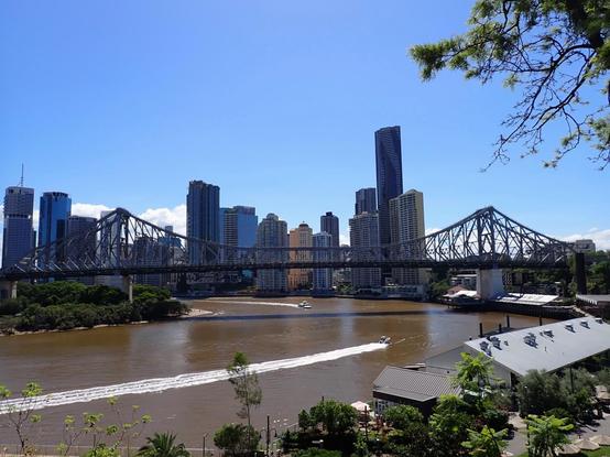 View of a suspension bridge spanning a river with tall buildings behind framed by a clear blue sky