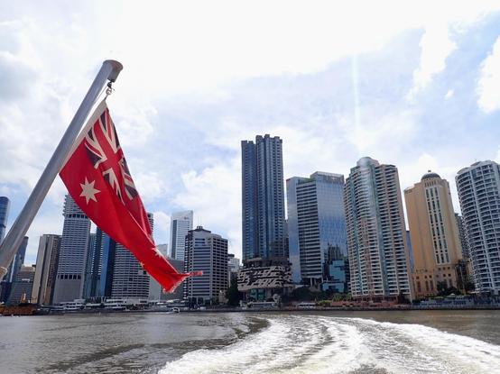 View of Brisbane skyline of tall buildings under a blue sky taken from a boat with Australian Red ensign flag flying in front