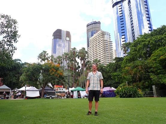Guy in checked shirt standing in grass park  in front of market stalls and tall buildings