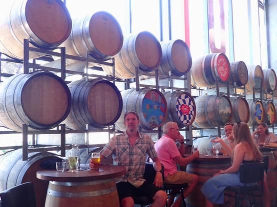 Guy with beer sitting at a barrel table in front of racked beer barrels in a large beer hall