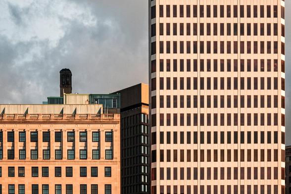 A color photograph showing three city buildings - one lower extending across almost half the frame from the left, a taller one on the right that rises out of the frame, both squarely facing the camera. Behind, a medium height building sits at a different angle, visible in the narrow space between.

The warm sun of the late afternoon on the buildings contrasts with the grayish, cloudy skies behind.