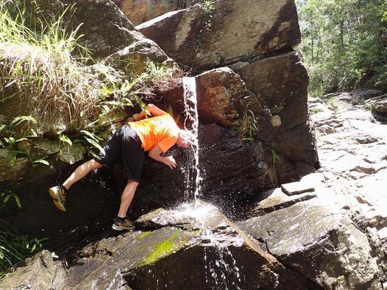 Guy in orange top dunking his head under a small trickle of water in a rocky outcrop
