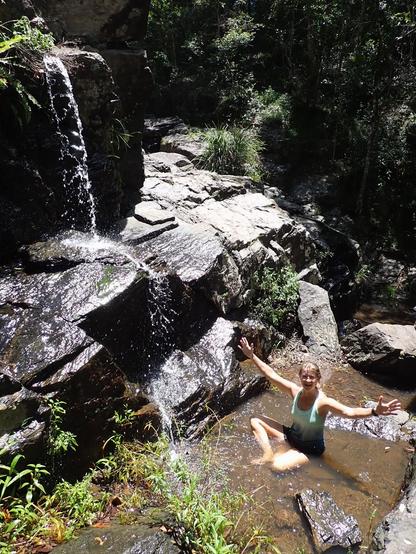 Lady in green vest top sitting in a shallow pool under a trickling waterfall from a stony outcrop