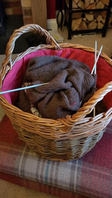 A wicker basket about as tall as it is round with two handles on the top edge. Lined in red fabric. It contains incomplete knitting in brown yarn with double pointed needles apparently shoved in at random.