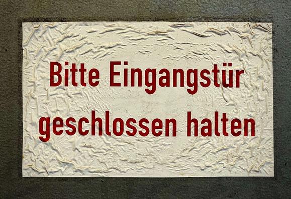 Gewelltes Schild mit der Aufschrift „Bitte Eingangstür geschlossen halten“