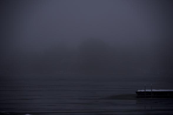 Foggy landscape with trees barely visible in the background and a snow-covered pier over a frozen lake in the foreground.