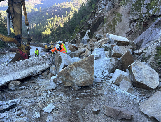 Personnel wearing orange and hard hats and yellow looking at very large rocks coming down a hillside, heavy equiment on left