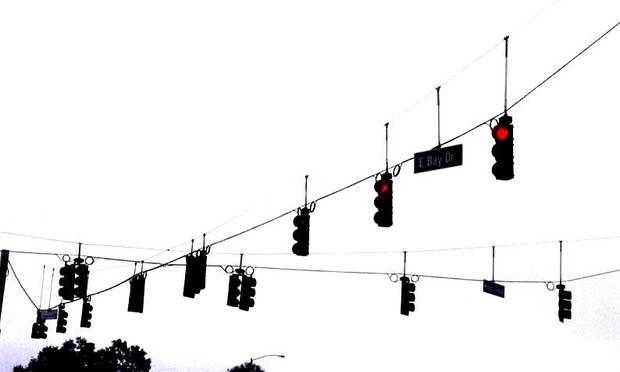Una docena de semáforos suspendidos de cables. En dos de ellos se ve que están en rojo. De fondo, solo el cielo blanco.