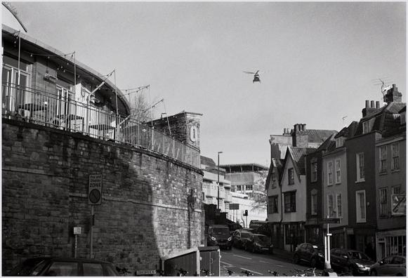 A black and white photograph of a Bristol, UK street scene.

On the left of the frame is a high stone wall on top of which is the Zero Degrees craft brewery and pub.
On the right are old house and business premises with vehicles parked outside and traffic going past.
In the gap between can be seen Bristol Royal Infirmary (B.R.I.) in winter sunlight.
In the sky at the top of the frame is a helicopter, that has just left the B.R.I