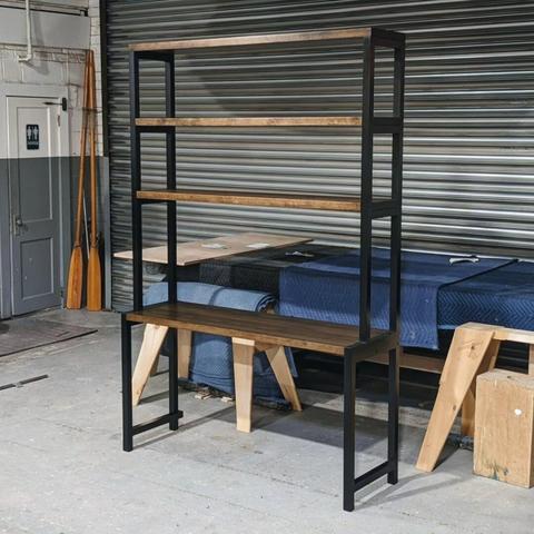 A combination desk and bookcase. A black frame holds a deep brown desktop with 3 shelves above it. The piece sits in the middle of a wood shop. Another piece of furniture is covered by blankets behind it.