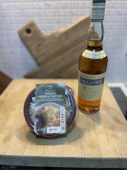 Ready made haggis with neeps and tatties alingside a bottle of  Cragganmore whisky. They are sitting on a butcher block and a wood cutting board in the shape of Texas is in the background.