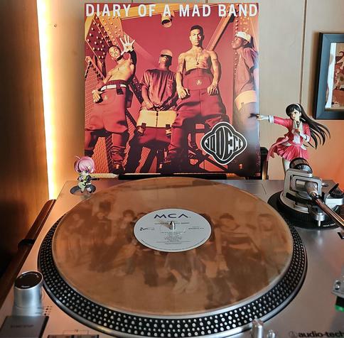 A Translucent Tan vinyl record sits on a turntable. Behind the turntable, a vinyl album outer sleeve is displayed. The front cover shows the 4 members of Jodeci posting around metal beams.