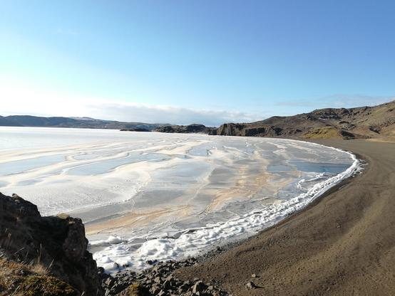 Une partie d'un lac totalement gelée dans un environnement rocailleux et dépourvu de neige. Le pourtour ressemble à de l’écume mais il s'agit de neige glacée. Le ciel est très dégagé.