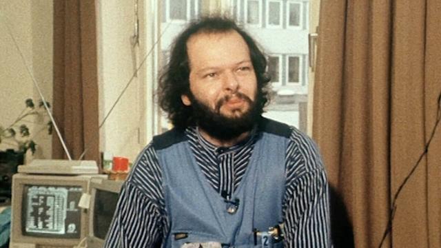 Das Bild zeigt einen Mann mittleren Alters mit vollem Bart und langen, welligen Haaren. Er trägt ein gestreiftes Hemd mit einer Latzhose und blickt direkt in die Kamera. Hinter ihm befindet sich eine ältere Computeranlage auf einem Tisch, im Hintergrund sind Vorhänge und ein Fenster sichtbar. Der Mann wirkt nachdenklich und entspannt. Er heißt Wau Holland und war ein toller Mann, der leider viel zu früh verstorben ist.