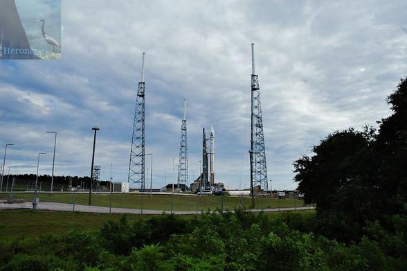 An outdoor, daylight, landscape photograph of Launch Complex 41 with an Atlas 5 rocket on the launchpad in the center. The rocket has a copper-colored first stage and white upper stage and conical fairing. Three truss towers are visible around the rocket.