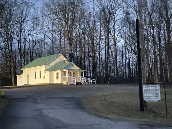 An old fashioned church building with low-roofed entryway and rectangular chapel with back addition for storage. Building has cream colored clapboards and a green roof. Stands against a backdrop of trees with a grassy front yard and a sign saying “Acorn Hill church of Christ”
All bathed I. Early morning light with a suspicion of blue sky through the trees