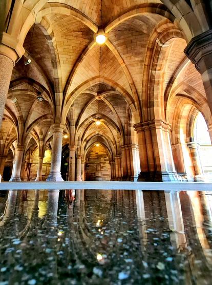 An area of cloisters with a gothic arched ceiling reflected in a puddle on the floor. In the reflection, there appears to be a ghost leaning on one of the columns.