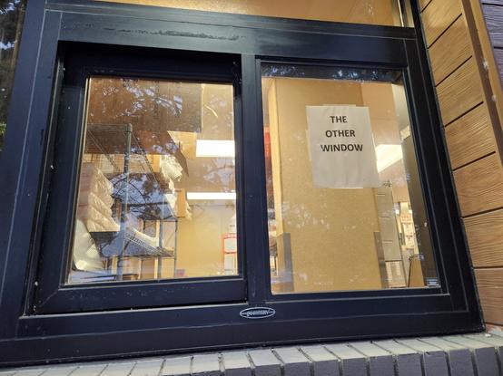 A fast food drive-thru window. Behind the glass, there is no cash register, no worker, and no indication that the window station is a working drive-thru station. On the left side behind the glass, there are some shelves of fast food restaurant supplies. 

On the right side of the glass, someone has hung a piece of paper that reads only "The Other Window."