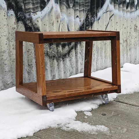 A white oak kitchen cart parked in a snowy spot on the sidewalk. The cart has two trapezoidal sides holding an upper and lower shelf. The cart is a warm brown color and has small, clear wheels.