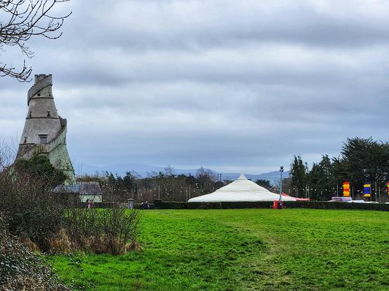 A view of the bottle shaped Wonderful Barn, the low wide bell shaped white pavilion where the Symposium is taking place, and banners in red and yellow, and blue and yellow marking where the food trucks are.