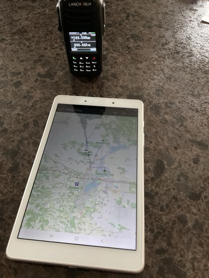 A HG-UV98 walkie-talkie and a tablet displaying an APRSdroid map are placed on a dark countertop.