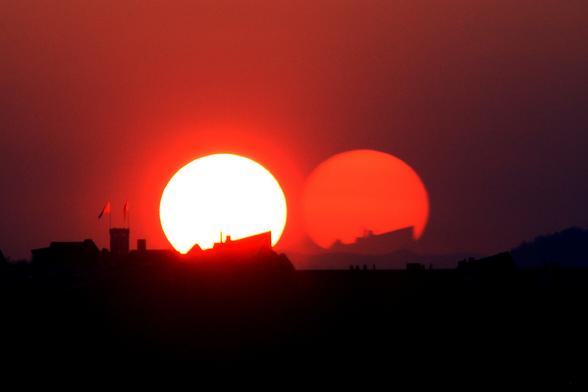 A photo of a sunset taken straight into the Sun, setting near the city castle and projecting the second, phantom Sun and a silhouette of a residential building's rooftop side by side with the real Sun onto the double pane window the photo was taken through.