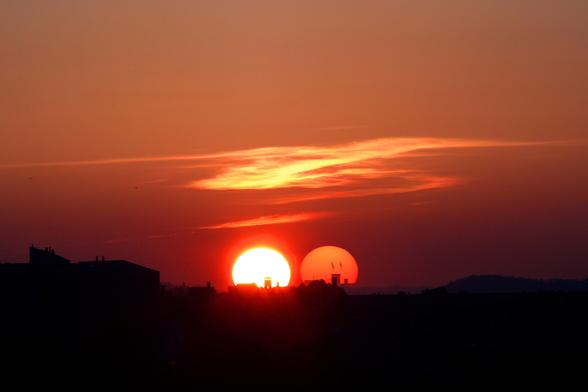 A photo of a sunset taken straight into the Sun, setting behind the city castle and projecting the second, phantom Sun and the castle tower silhouette side by side with the real one onto the double pane window the photo was taken through.
