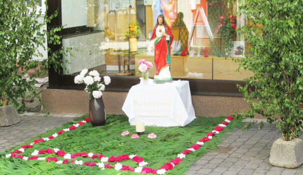 Vor einem bunten Schaufenster steht eine bunte Jesusfigur auf
einem kleinen, leintuchumhüllten Podest, darunter ganz viel
Grünstreu und Blumen.