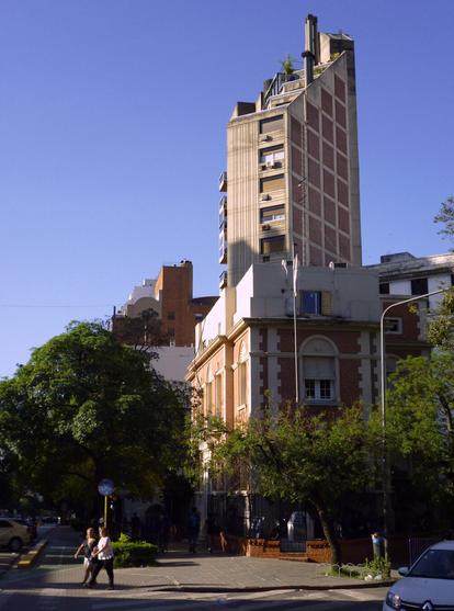 A city street corner with a mix of modern and traditional architecture, surrounded by green trees, with pedestrians walking, and a car crossing the road on the right bottom corner. The sunlight causes some hard contrast of light and shadows.
