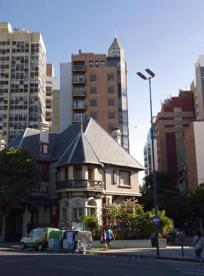 An eclectic urban street scene with a Victorian-style house with a garden with green plants and red flowers in the foreground flanked by modern high-rise buildings, streetlights, pedestrians, and a parked vehicle with advertising.