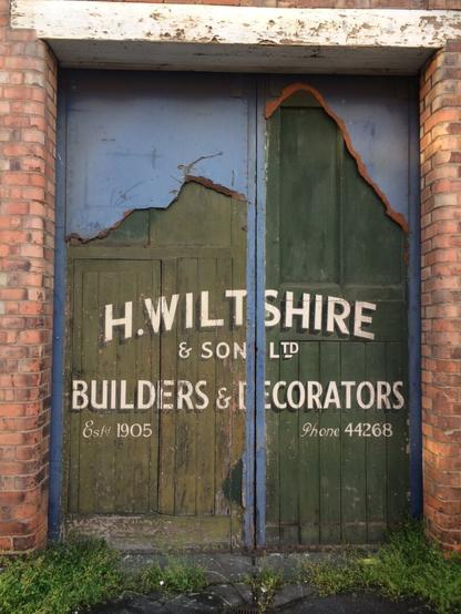 Double doors to a red brick building with a white lintel above. They are painted green, in poor condition, with a blue laminate coming away to reveal faded white lettering with a block shade that reads: "H. Wiltshire & Sons Ltd, Builders & Decorators, Est. 1905, Phone 44286".